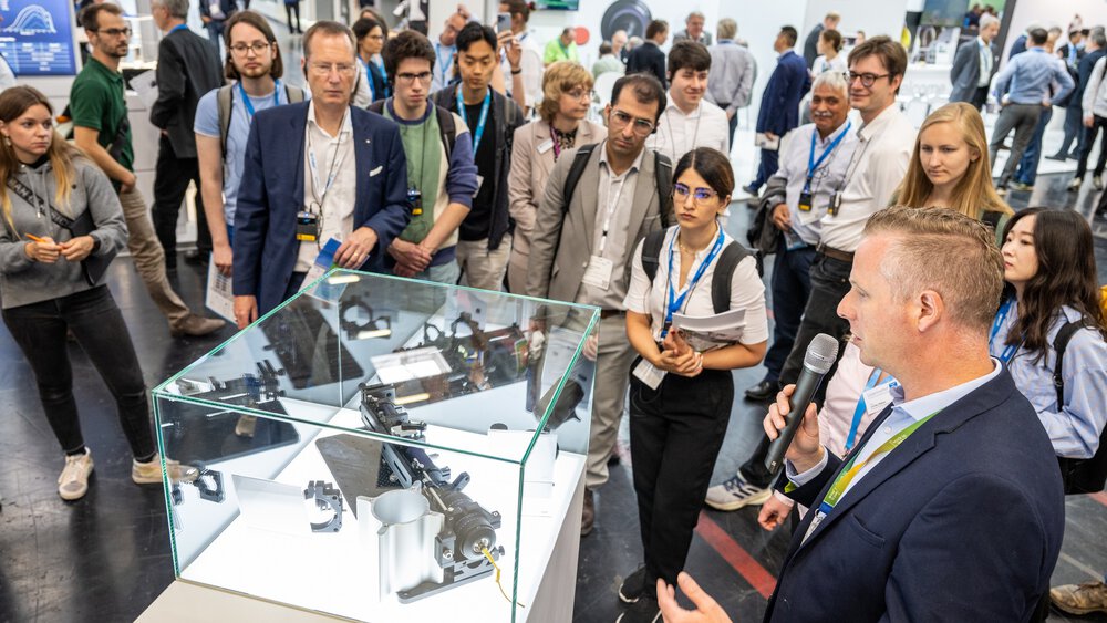 Eine Gruppe von Menschen versammelt sich um eine Glasvitrine, während ein Mann mit einem Mikrofon eine Präsentation hält.