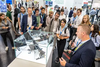 Eine Gruppe von Menschen versammelt sich um eine Glasvitrine, während ein Mann mit einem Mikrofon eine Präsentation hält.