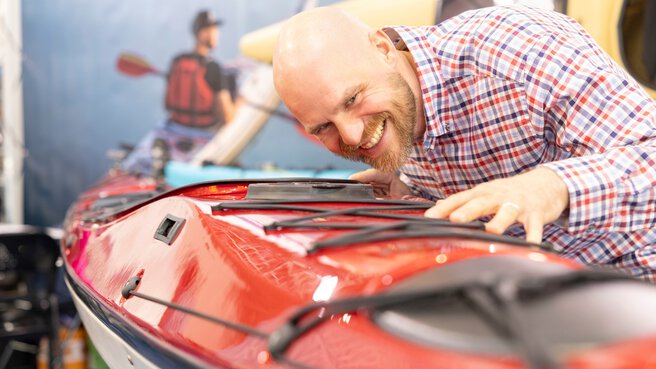 A man leans over a kayak on display and looks at it closely with a smile. In the background, there is a trade fair wall with a water sports motif.