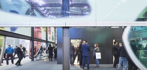 Blick durch eine Wand aus bunten abstrakten Fensterfronten, hinter der sich mehrere Gruppen von Messebesuchern in Businesskleidung unterhalten.