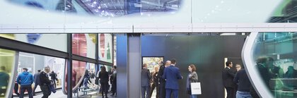 Blick durch eine Wand aus bunten abstrakten Fensterfronten, hinter der sich mehrere Gruppen von Messebesuchern in Businesskleidung unterhalten.