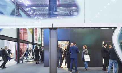 Blick durch eine Wand aus bunten abstrakten Fensterfronten, hinter der sich mehrere Gruppen von Messebesuchern in Businesskleidung unterhalten.