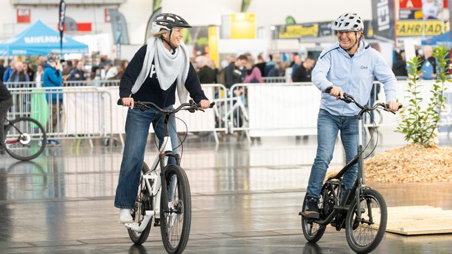 A man and a woman wearing helmets sit laughing on bicycles and test them on a separate area at f.re.e.