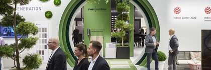 A white wall with a circular, green stepped passageway with hanging plants through which other exhibition stands can be seen. People walk past in the foreground.