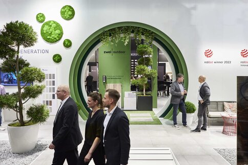 A white wall with a circular, green stepped passageway with hanging plants through which other exhibition stands can be seen. People walk past in the foreground.