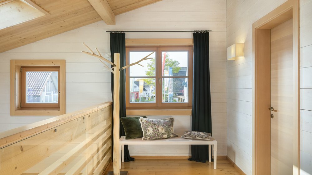 Gemütlicher Flur mit Holzdecke, Sitzbank am Fenster, Kissen und schwarzen Vorhängen. Helles Tageslicht erhellt den Raum.