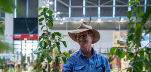  Mann mit Hut steht an einem Messestand zwischen jungen Bäumen und betrachtet eine Pflanze in einer Ausstellungshalle.