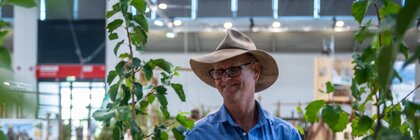 A man wearing a hat stands at an exhibition stand between young trees and looks at a plant in an exhibition hall.