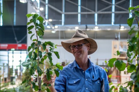 A man wearing a hat stands at an exhibition stand between young trees and looks at a plant in an exhibition hall.