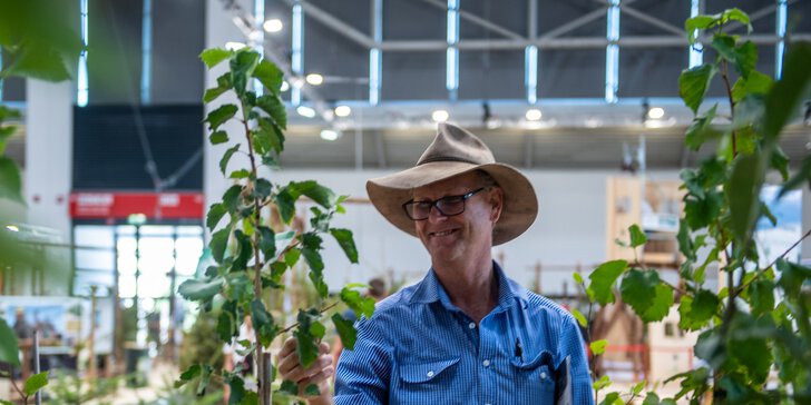 Mann mit Hut steht an einem Messestand zwischen jungen Bäumen und betrachtet eine Pflanze in einer Ausstellungshalle.