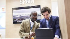 Two men stand at an exhibition booth labeled “Spain” at EXPO REAL in Munich. They are looking at a tablet together, discussing something, while one of them also holds a smartphone. In the background, a screen displays a presentation about available industrial land in Spain. On the table, there is a small rubber duck.