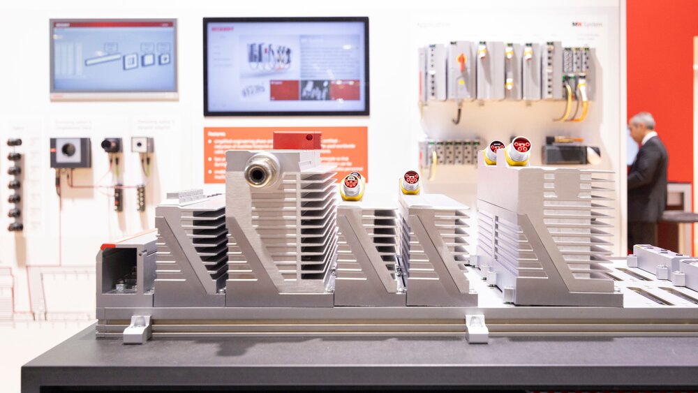 Eine Messestand auf der productronica mit mehreren Bildschirmen und Exponaten. An der Wand steht „MX-System: modular and pluggable“.