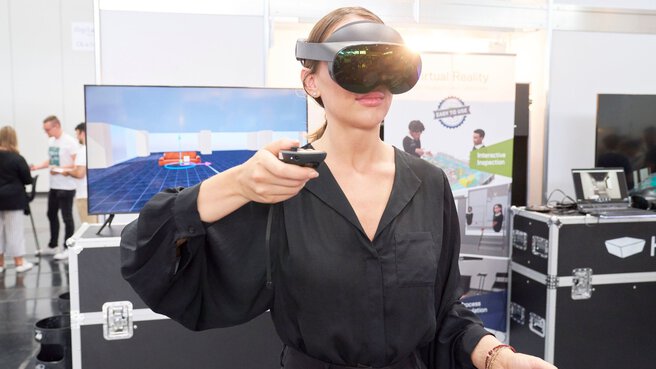 Person using a virtual reality headset and controller at a tech event, with your view displayed on a screen in the background.