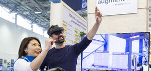 Ein Besucher auf einem Messestand trägt ein Augmented-Reality-Headset, eine Ausstellerin steht neben ihm und demonstriert die Handhabung.