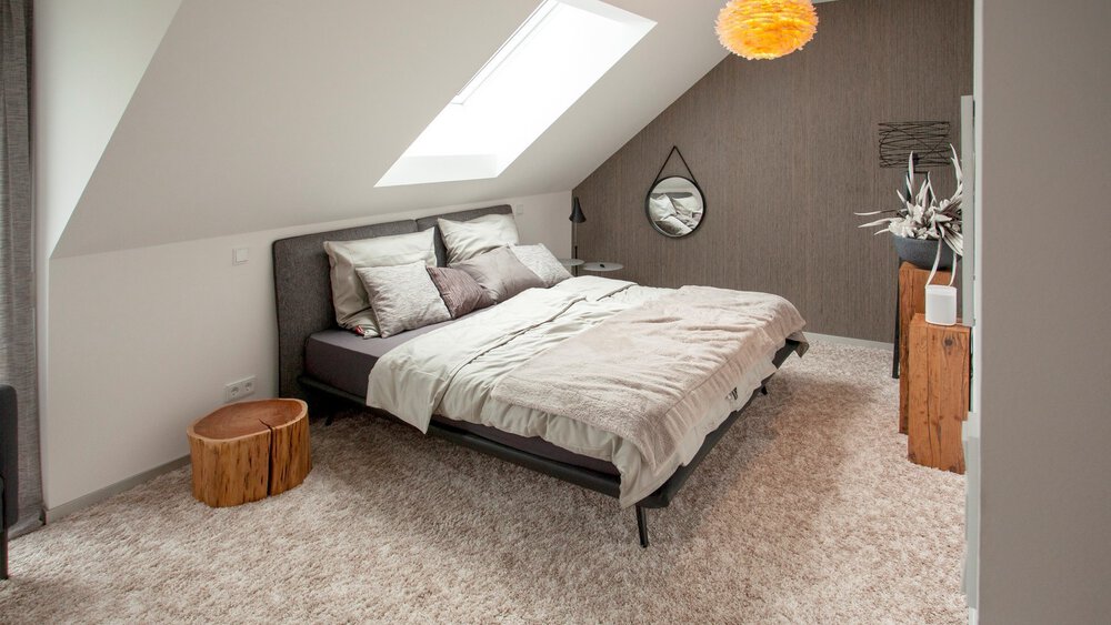 Ein gemütliches Schlafzimmer im Dachgeschoss mit Oberlicht, dunklem Bettgestell, grauer Akzentwand und Holzkommode.