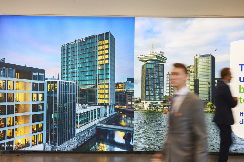 Two men in suits walk past a large billboard with city views of modern buildings in Hamburg and Amsterdam.