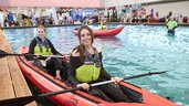 Zwei Frauen sitzen in einem Kajak, welches sich auf einem großen Indoor-Pool in einer Messehalle befindet und lachen in die Kamera.