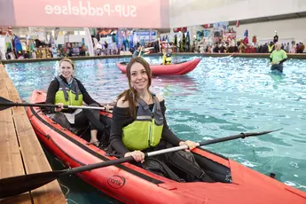 Two women sit in a kayak on a large indoor pool in an exhibition hall and smile at the camera.