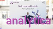 3D letters in bright pink forming the word 'analytica'. Behind it, a visitor stands in front of a poster with 
