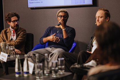 Four people sit on a podium in front of an audience with microphones and talk to each other.