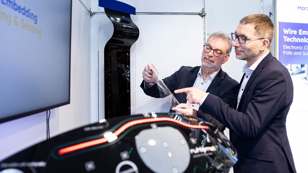 Two professionals discussing flexible electronics at a booth with an automotive demonstrator in the foreground