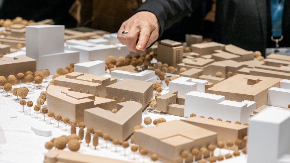 A hand points to a building in an architectural model of a city with buildings and trees.