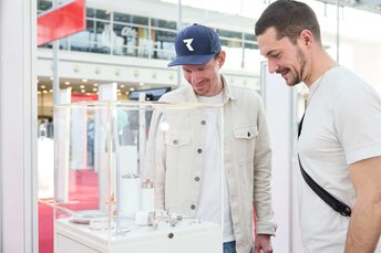 Zwei Messebesucher betrachten interessiert ein Exponat in einer Vitrine.