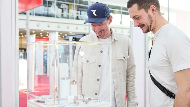 Two trade fair visitors examine an exhibit displayed in a glass case.