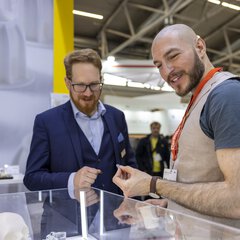 Two men smile and interact at an exhibition stand, one holding a 3D-printed object.