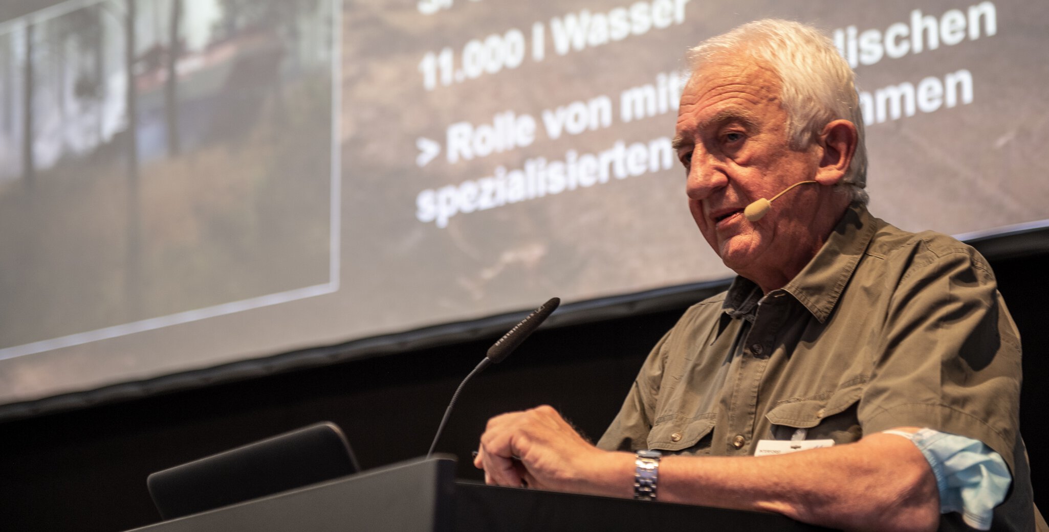 Elderly man speaking at a podium with a presentation screen in the background at INTERFORST.