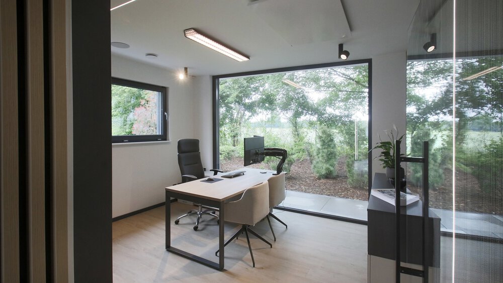 Modernes Büro mit großen Fenstern, zwei Stühlen, einem Schreibtisch und Blick ins Grüne.