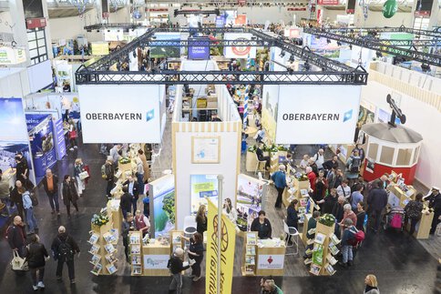 Überblick über eine belebte Messehalle mit zahlreichen Messeständen und Besuchern, zentraler 'Oberbayern'-Stand, Informationsdisplays und Gängen voller Menschen.