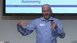A man in a blue shirt is giving a presentation and gesticulating with his free hand.