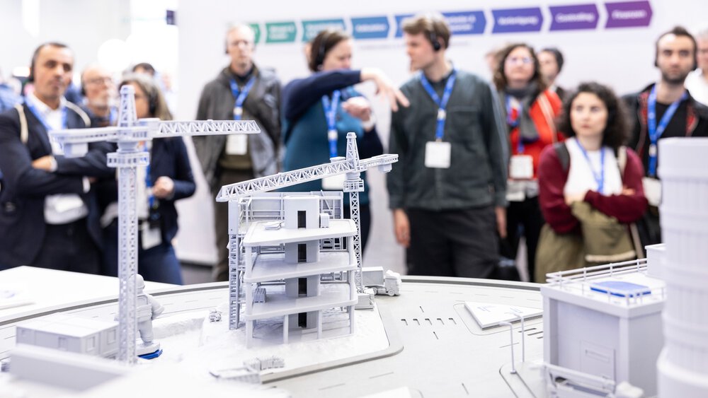 Eine Gruppe von Menschen betrachtet auf einer Ausstellung eine Modellbaustelle mit Kränen und Gebäuden.