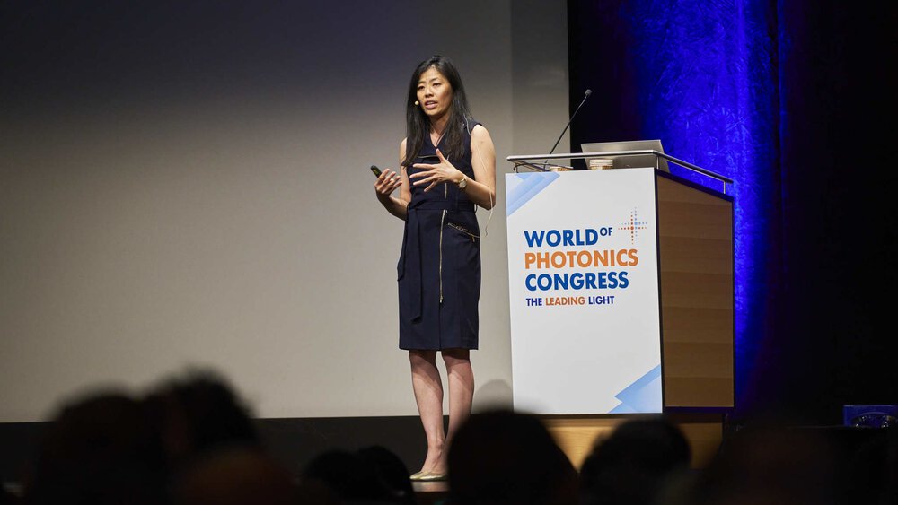 A woman speaks on the stage of the World of Photonics Congress, the event logo can be seen on the podium next to her.