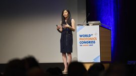 A woman speaks on the stage of the World of Photonics Congress, the event logo can be seen on the podium next to her.