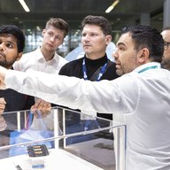 An exhibitor shows a group of four interested men something on a display above a glass case.