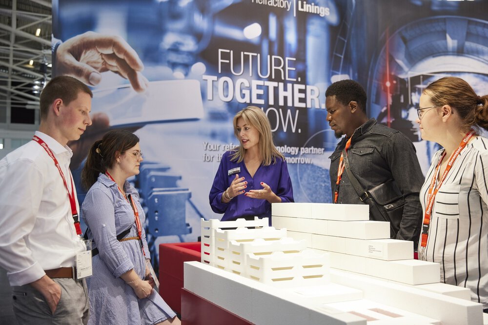 Eine Ausstellerin mit blonden Haaren spricht mit vier Messebesuchern an einem Stand auf der ceramitec. Zwischen ihnen liegen weiße gestapelte Ausstellungsstücke.