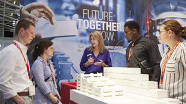 Eine Ausstellerin mit blonden Haaren spricht mit vier Messebesuchern an einem Stand auf der ceramitec. Zwischen ihnen liegen weiße gestapelte Ausstellungsstücke.