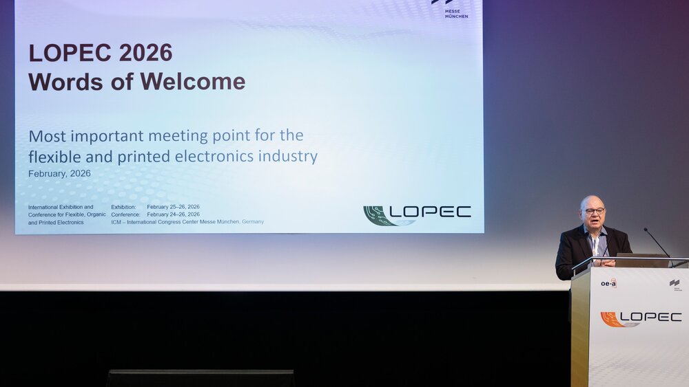A man in a suit is standing at a lectern in front of a large screen showing a presentation. The words “LOPEC 2026 Words of Welcome” are displayed on the screen.