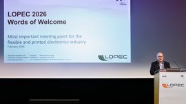 A man in a suit is standing at a lectern in front of a large screen showing a presentation. The words “LOPEC 2026 Words of Welcome” are displayed on the screen.