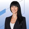 Frau mit langen schwarzen Haaren und schwarzem Blazer lächelt vor modernem, abstraktem Hintergrund in Blautönen und geometrischen Formen.