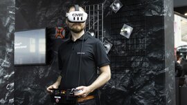 Mann mit VR-Brille und Steuergerät bei einer Technologie-Demonstration an einem Messestand mit dunklem Hintergrund.