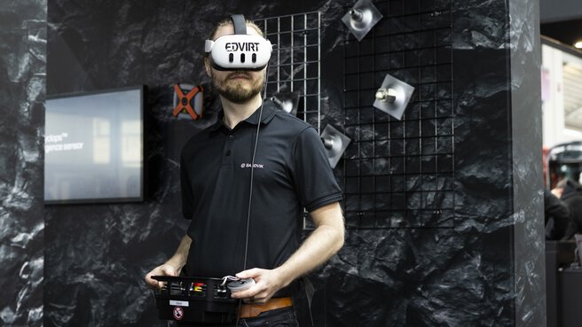 Man wearing a VR headset and controller during a tech demo at a trade fair booth with a dark backdrop.