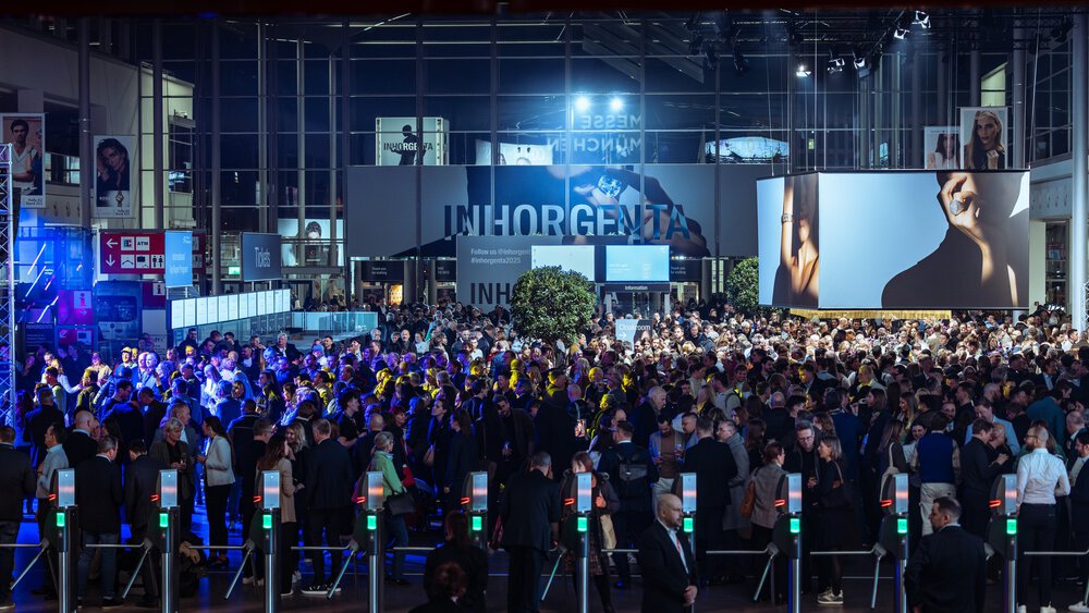 A large crowd gathers at the entrance to an indoor event with bright lighting and digital screens.