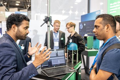 Two men talk at a technology stand where a laptop and devices are on display.