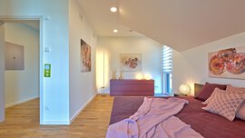 Ein helles Schlafzimmer mit rosa Bettwäsche, moderner Kunst an den Wänden und einer offenen Tür, die zu einem Flur führt.