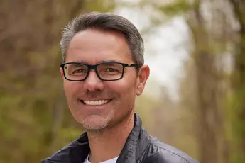 Ein lächelnder Mann, Tobias Gröber, mit Brille, einer schwarzen Jacke und einem weißen T-Shirt, steht auf einem Waldweg.