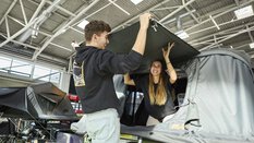 A man and a woman set up a roof tent together on a car at a trade fair and laugh.