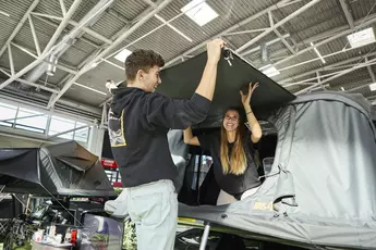 Ein Mann und eine Frau bauen gemeinsam ein Dachzelt auf einem Auto auf einer Messe auf und lachen dabei.